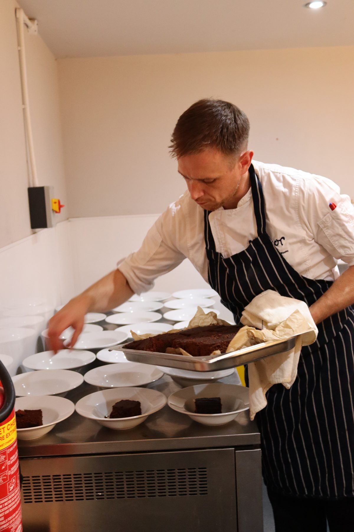 A chef in Manor House whites and a striped apron lifting a tray of warm chocolate brownie from the oven and portioning it into rows of white bowls laid out across the pass