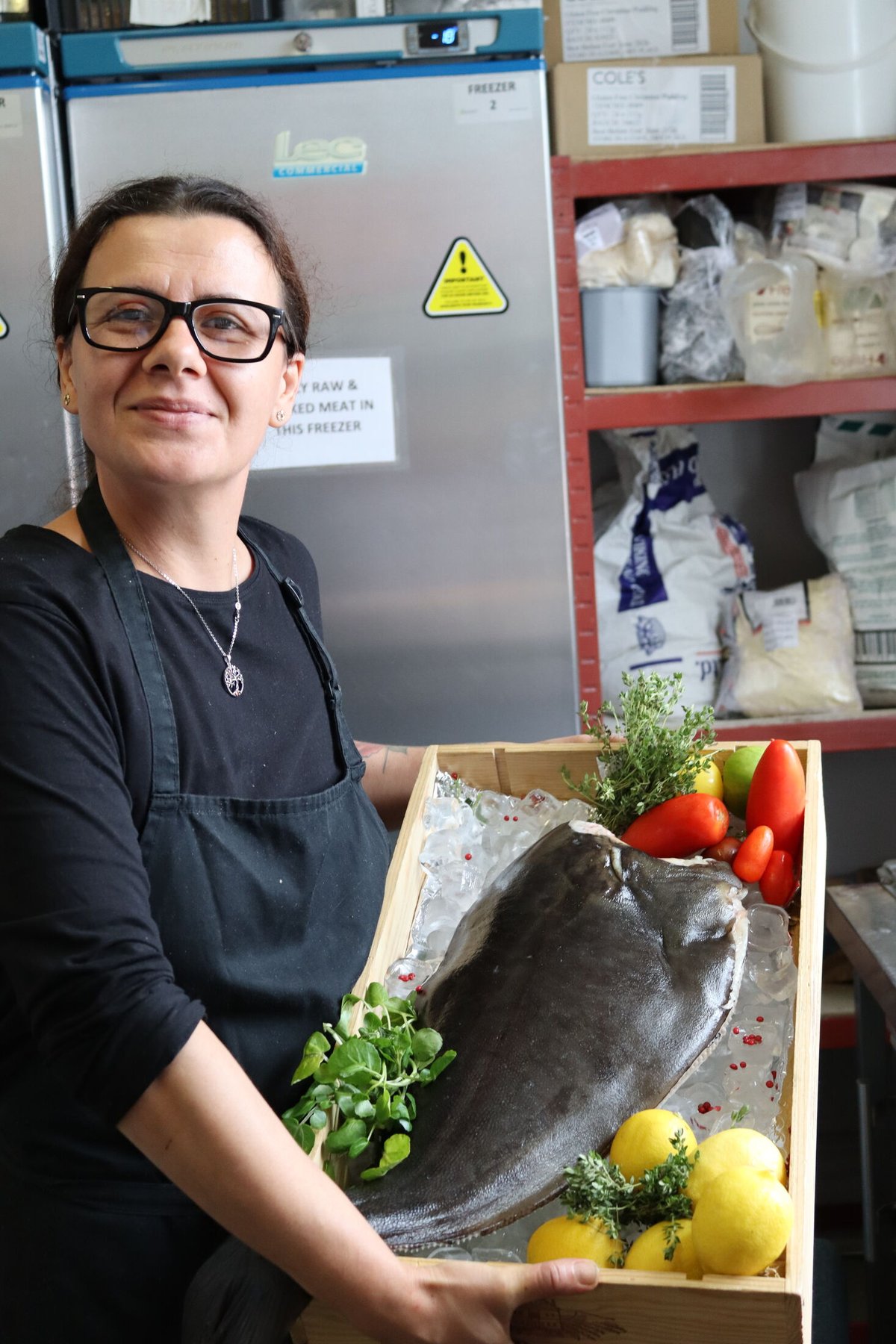 A chef in the kitchen holding a wooden crate of ice with a whole turbot laid on it, dressed with lemons, tomatoes, watercress and thyme