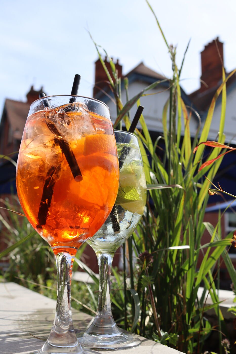 A tall orange spritz filled with ice and citrus, photographed close up among ornamental grasses with rooftops beyond