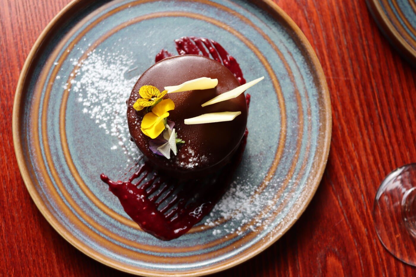 An overhead view of a domed chocolate dessert dusted with icing sugar, finished with a yellow flower and a smear of dark red fruit sauce
