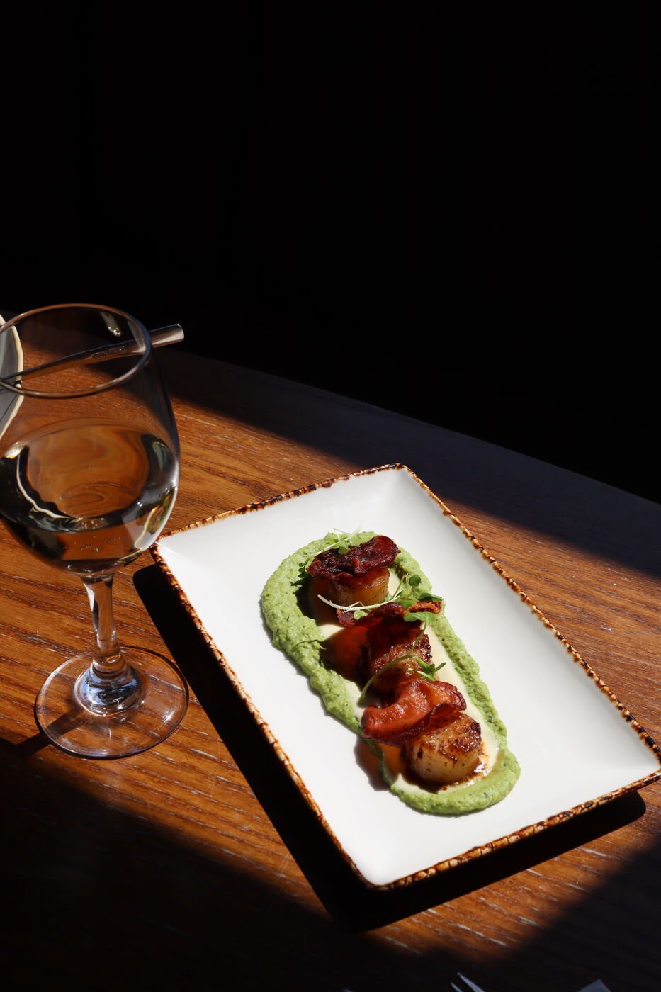 Three seared scallops with crisp bacon, set in a ring of bright green pea purée on a long white plate, lit by a shaft of low sunlight beside a glass of white wine