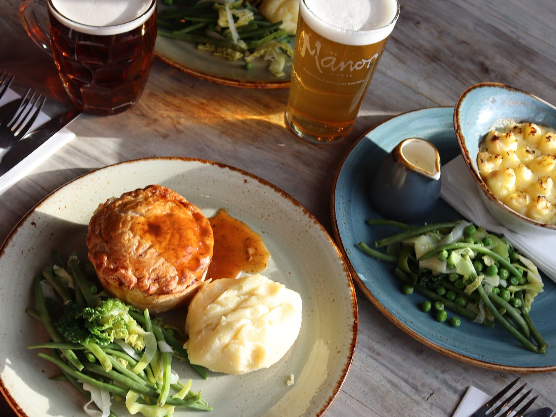 A golden pastry-topped pie with mashed potato, greens and gravy, served alongside macaroni cheese, a pint of ale and a pint of lager in a branded Manor House glass