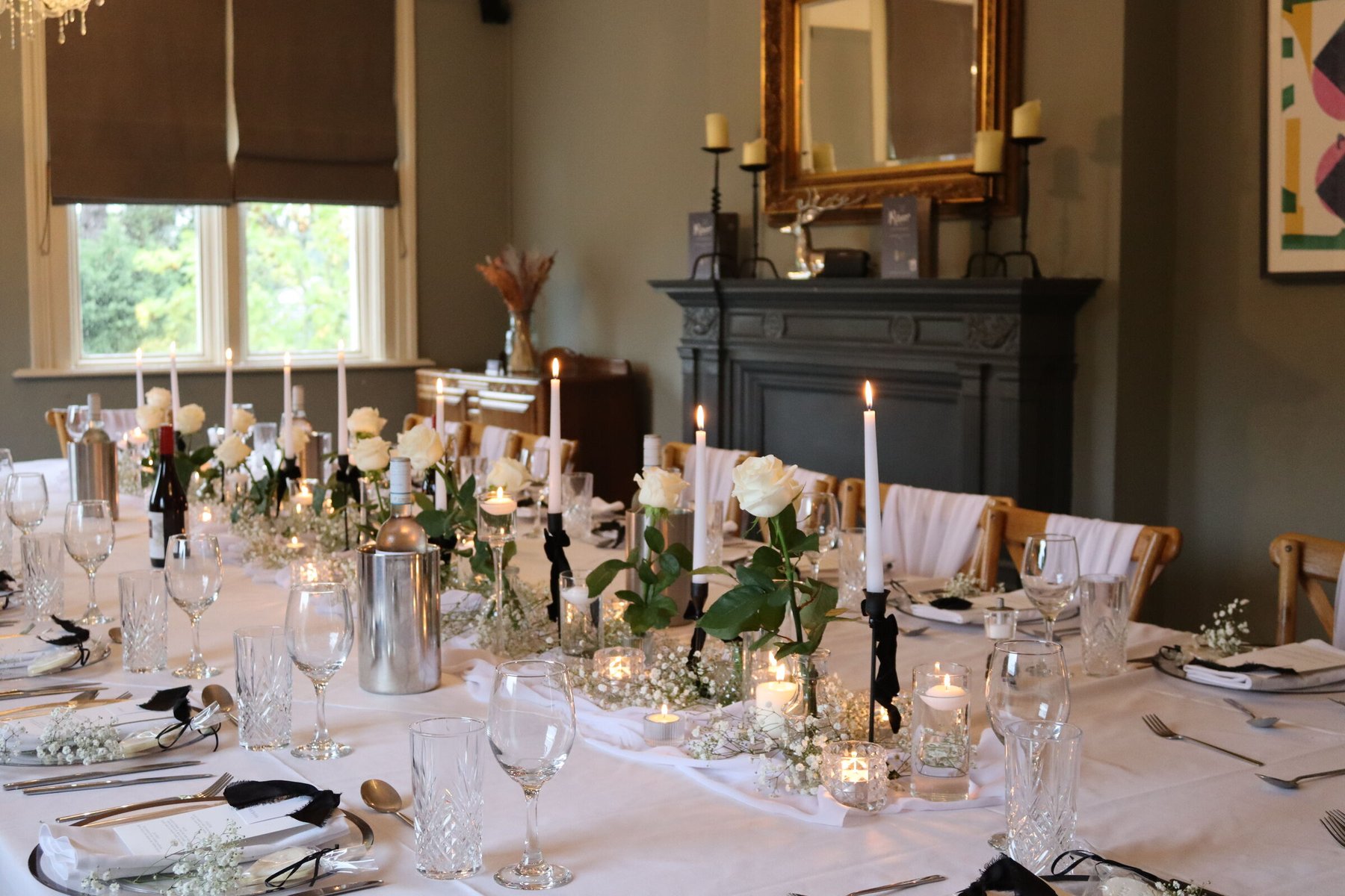 The private dining room dressed for a celebration — a long table in white linen with tall candles, white roses and gypsophila down its length, wine chilling in ice buckets, a dark carved fireplace and gilt mirror behind