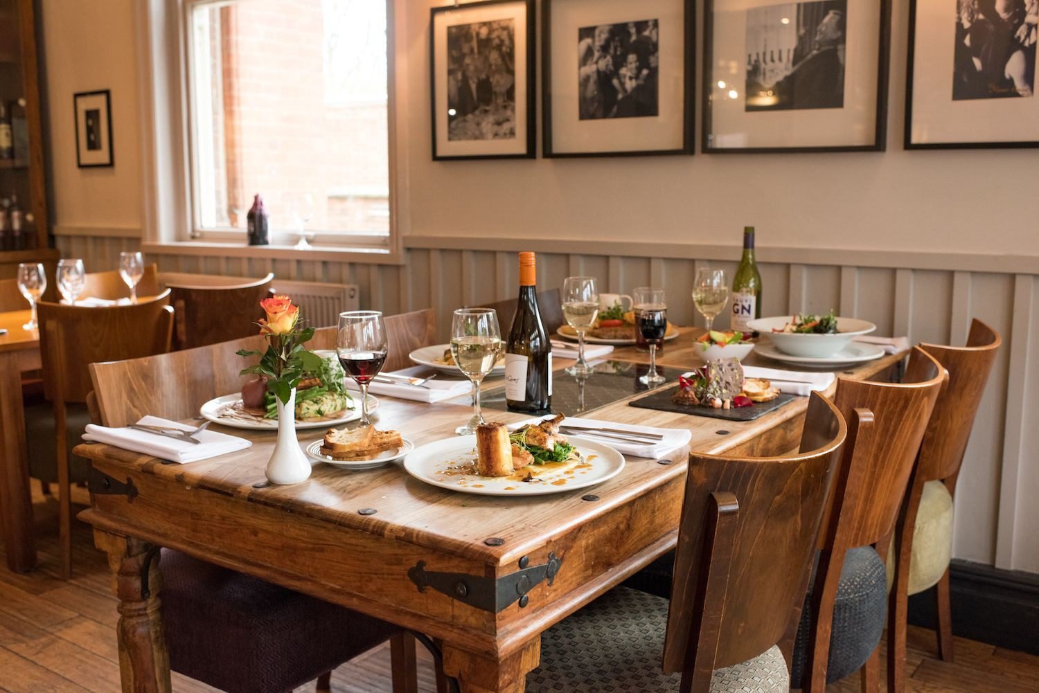 A heavy timber table in the restaurant laid with plated dishes, wine glasses and bottles, a single rose in a vase, with framed black and white photographs on the wall behind