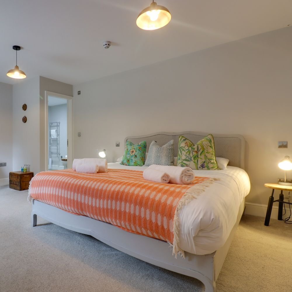 A bedroom with a cane headboard, a coral geometric throw and tropical print cushions, with an ensuite visible through the open door