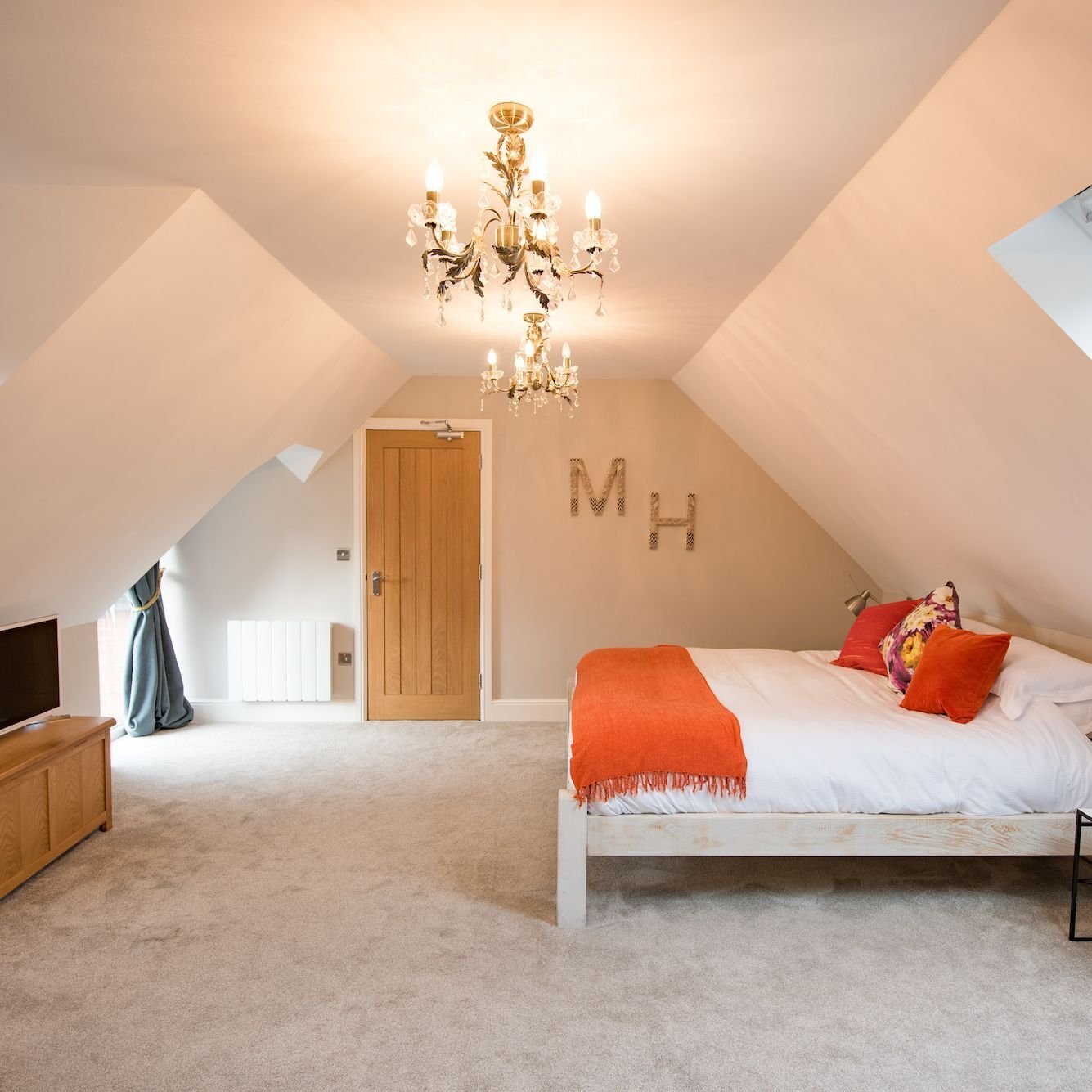 A bedroom set under twin eaves with a crystal chandelier, an orange throw across the bed and metal M and H letters on the wall