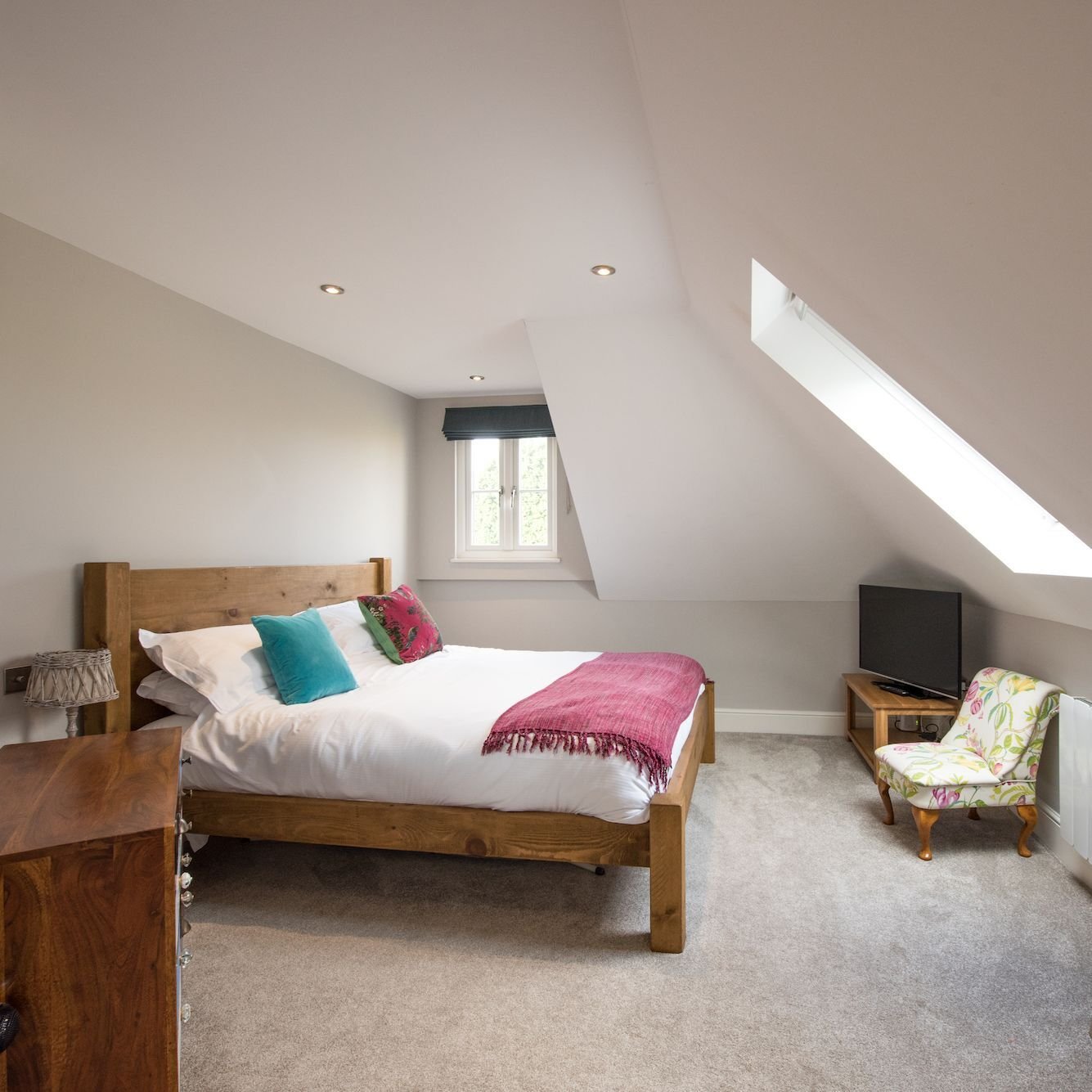 A bedroom under the eaves with a skylight, a solid timber bed frame, a raspberry throw and teal cushions, and a floral armchair in the corner