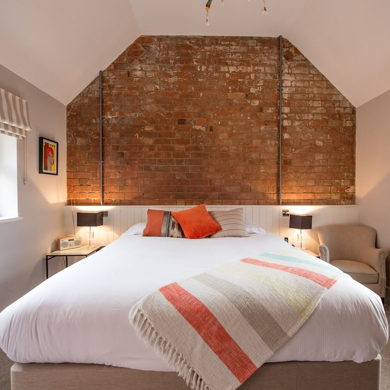 A bedroom with a full exposed brick gable behind the bed, white panelling below it, orange cushions and a striped throw, with reading lamps either side