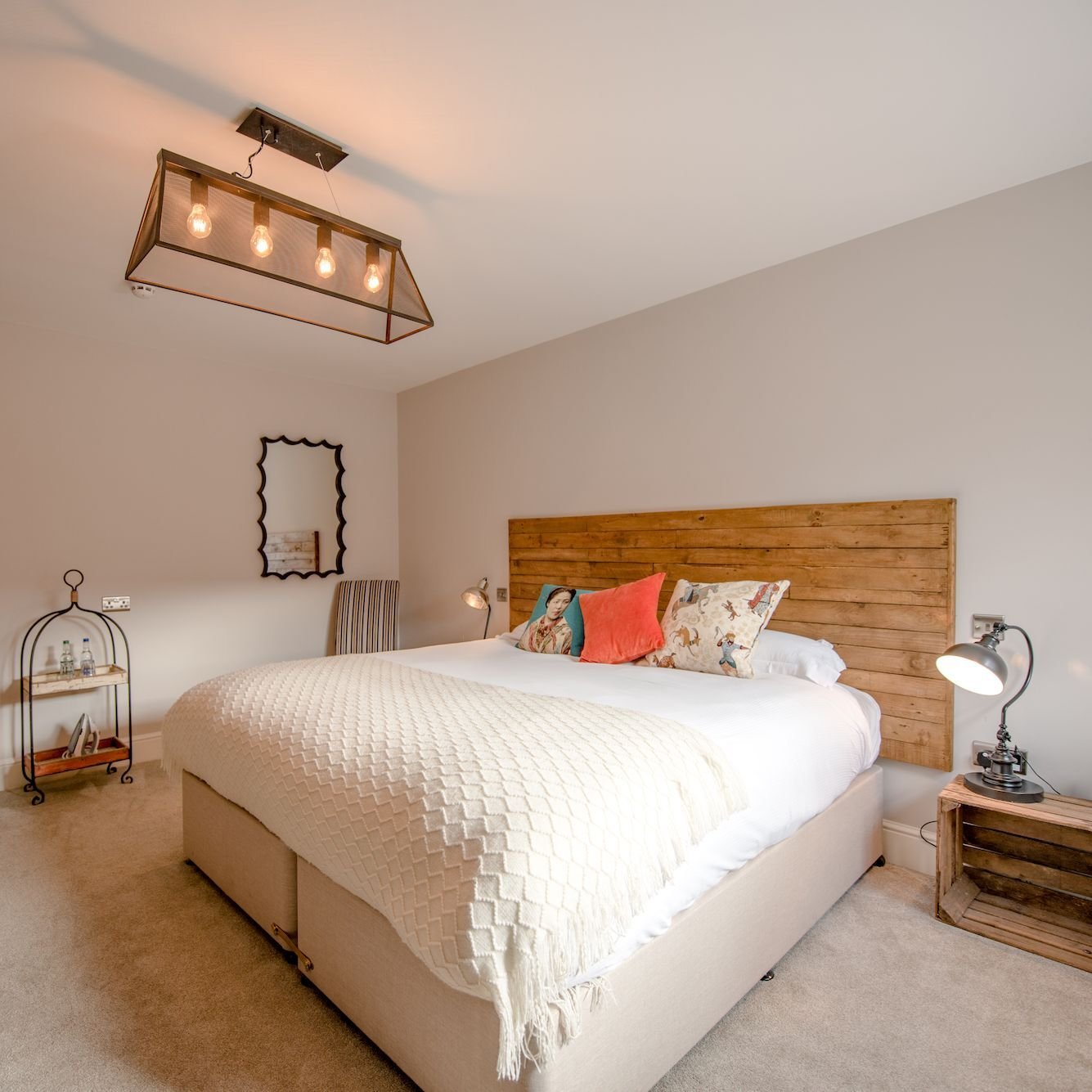 A bedroom with a wide reclaimed timber headboard, a coral cushion, an industrial glass pendant light and a wooden crate as a bedside table