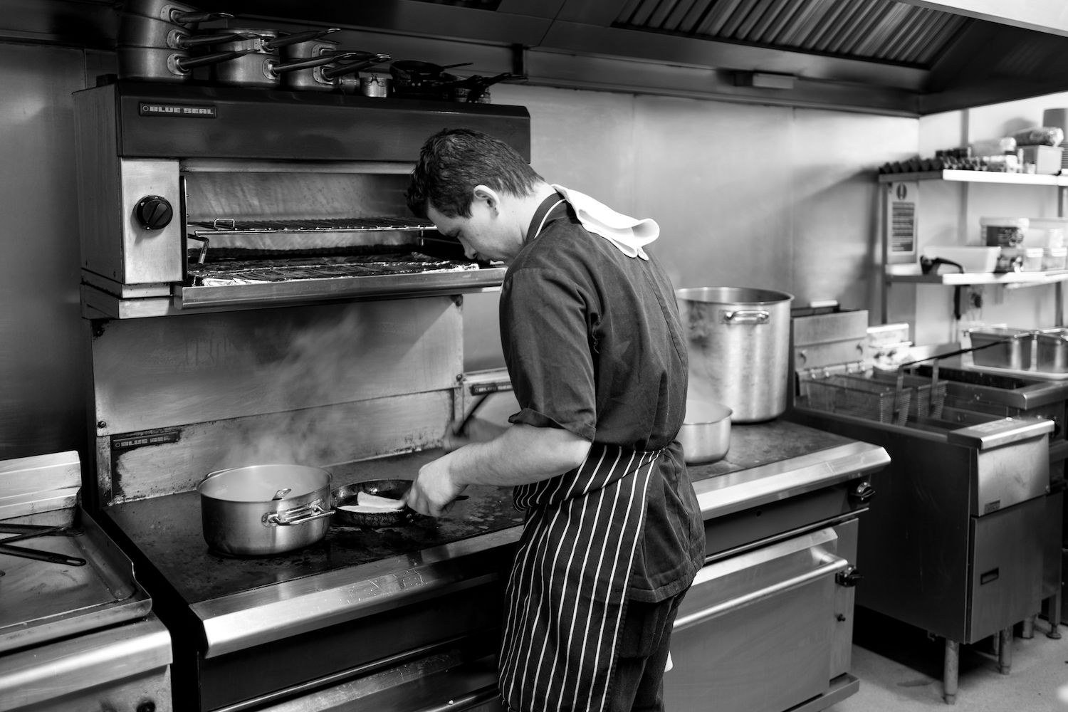 A chef in a striped apron working at the stove, photographed in black and white, pans going beneath the extraction hood