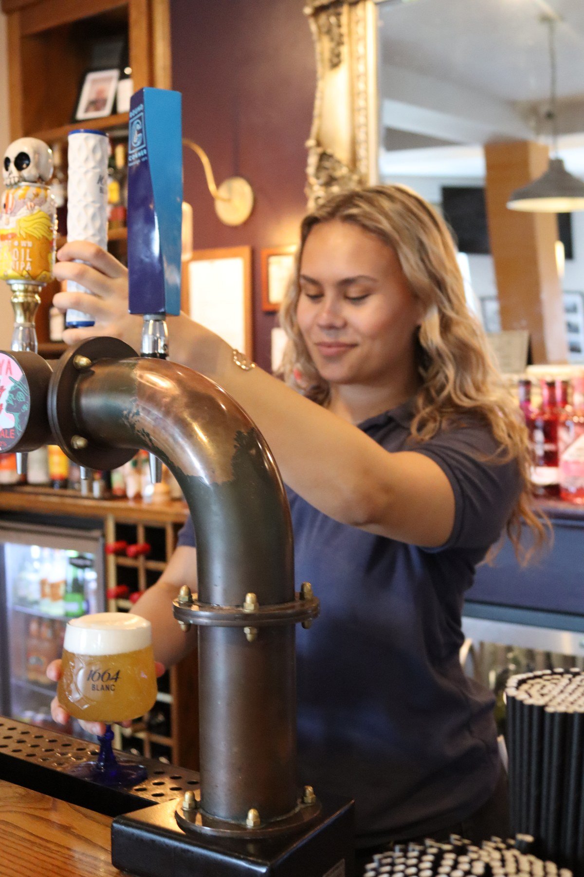 A member of the bar team pulling a pint at the hand pumps, tilting the glass under the tap, the bar and its optics behind her