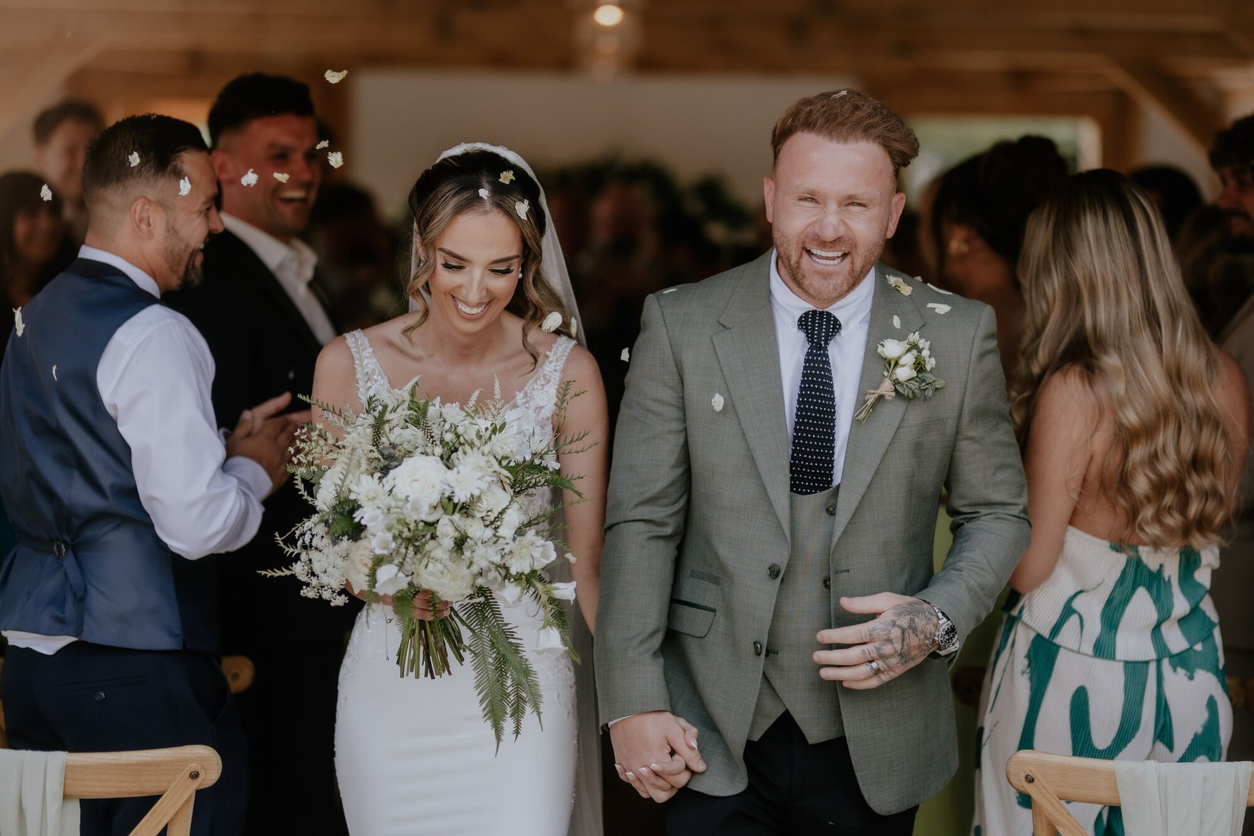 A newly married couple walking back down the aisle hand in hand, laughing, as guests throw white petals