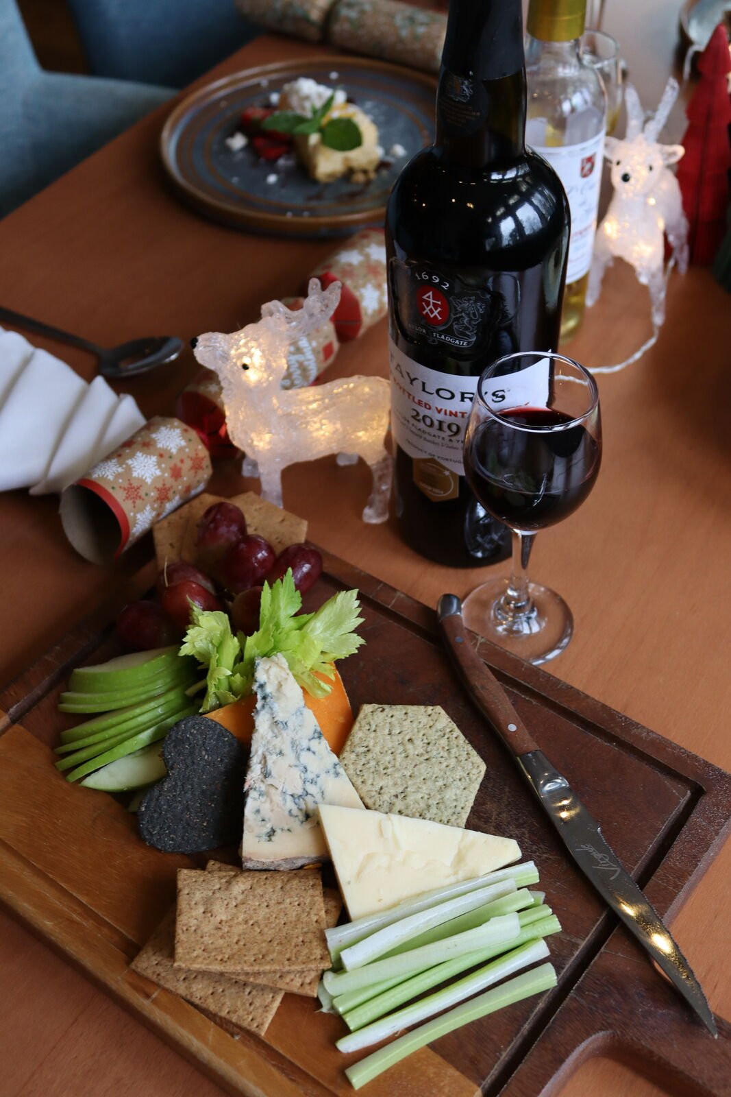 A cheeseboard with blue cheese, cheddar, red leicester, crackers, celery, apple and grapes, with a bottle of Taylor's 2019 port, a filled glass and festive crackers alongside