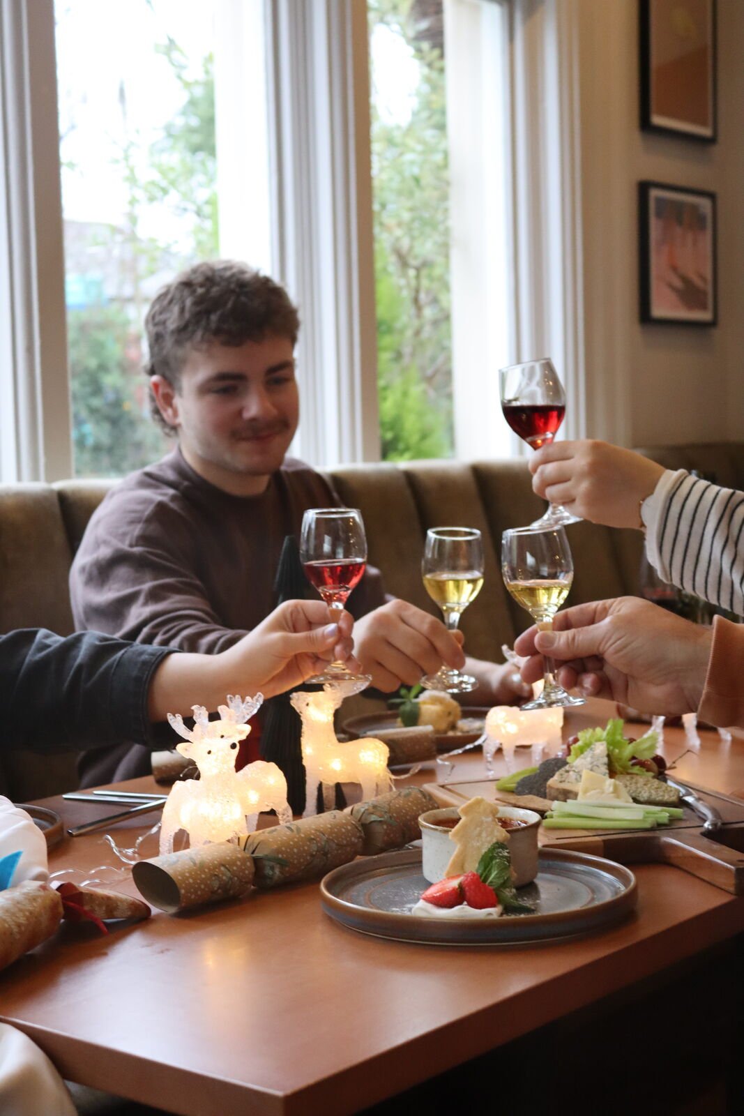 Guests raising glasses of dessert wine across a table set with crackers, desserts and light-up reindeer, daylight through the windows behind